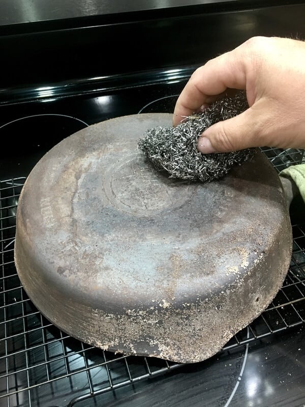 man's hand rubbing dirty cast iron skillet with steel wool