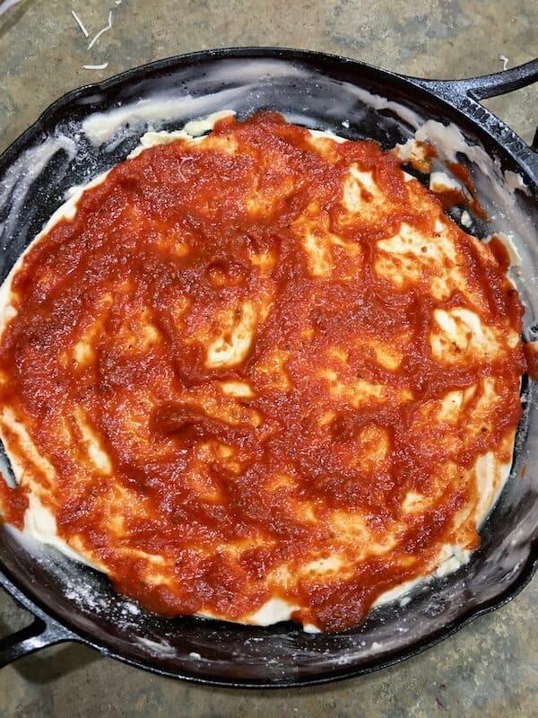 pizza sauce spread on top of pizza dough in a cast iron skillet