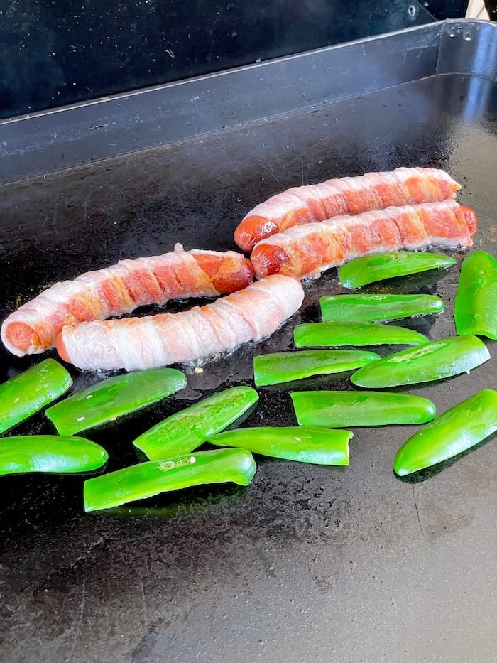 bacon wrapped hot dogs and jalapenos cooking on a griddle