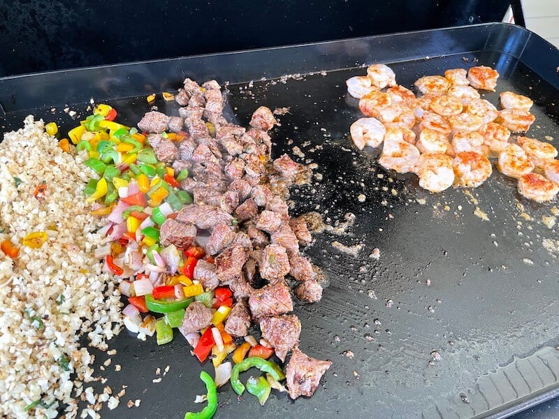 cauliflower rice, vegetables, steak, and shrimp cooking on a flat top grill