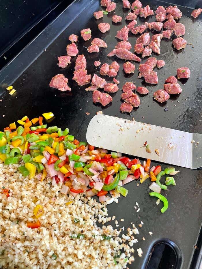 cauliflower rice, vegetables, and steak cooking on a griddle