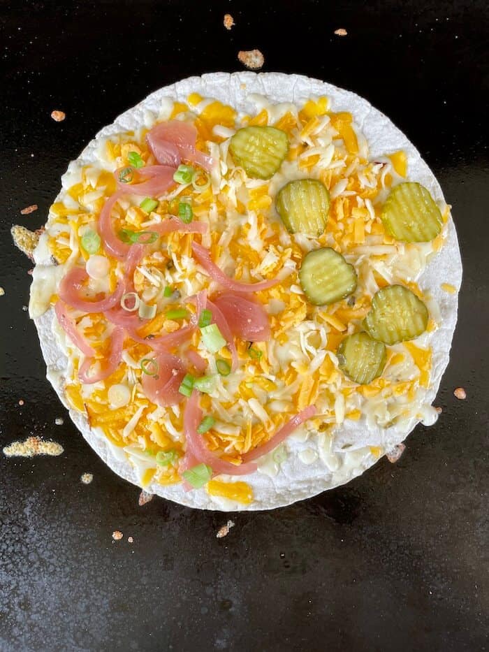 flour tortilla with shredded cheese, onions, and pickles cooking on a griddle