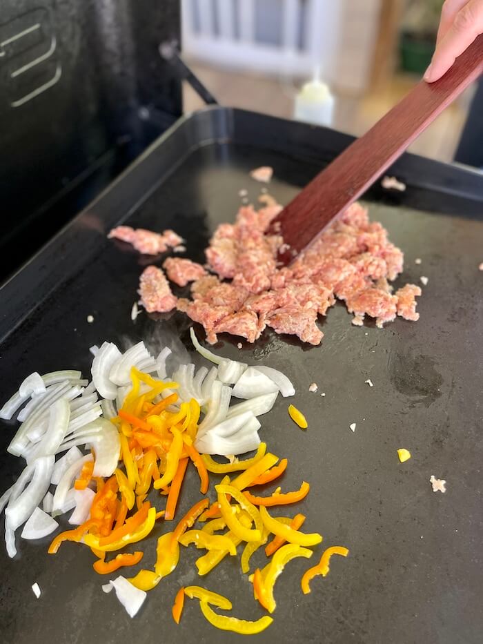 Italian sausage, bell peppers, and onions cooking on a griddle