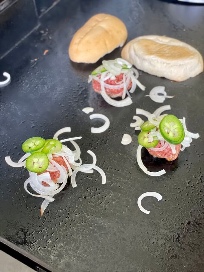 burger balls with sliced onions and jalapenos on a griddle