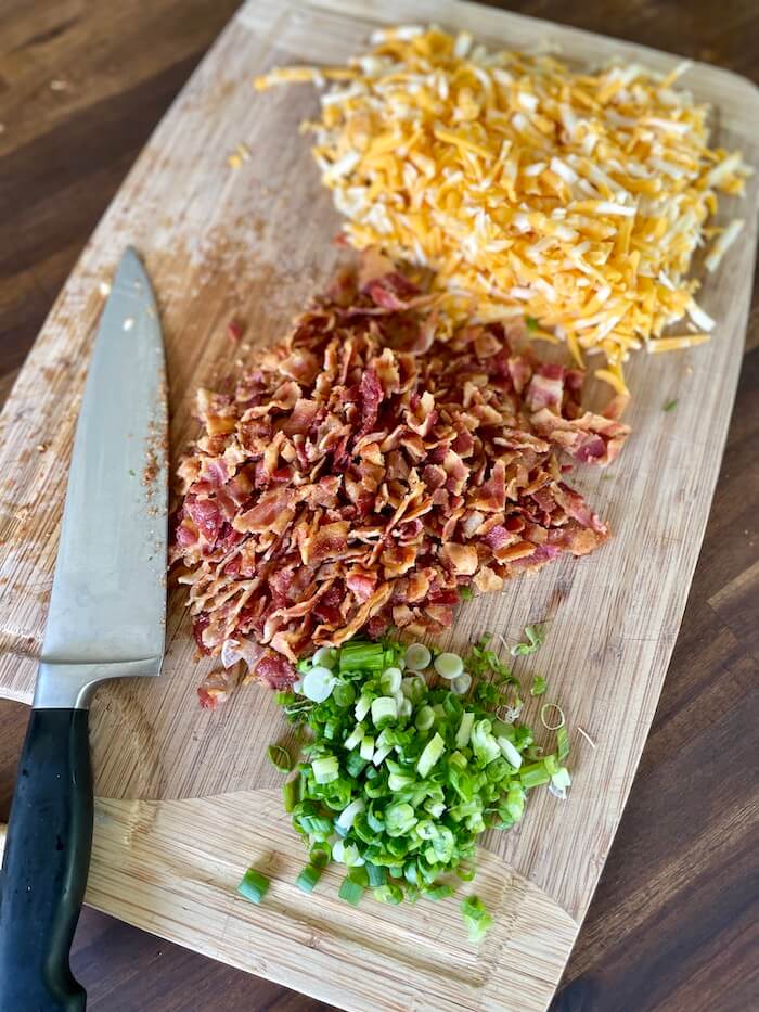 shredded cheese, chopped bacon, and sliced green onions on a cutting board