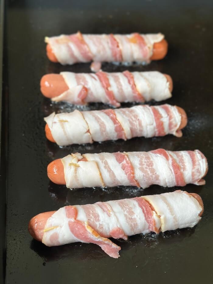 bacon wrapped hot dogs cooking on a griddle
