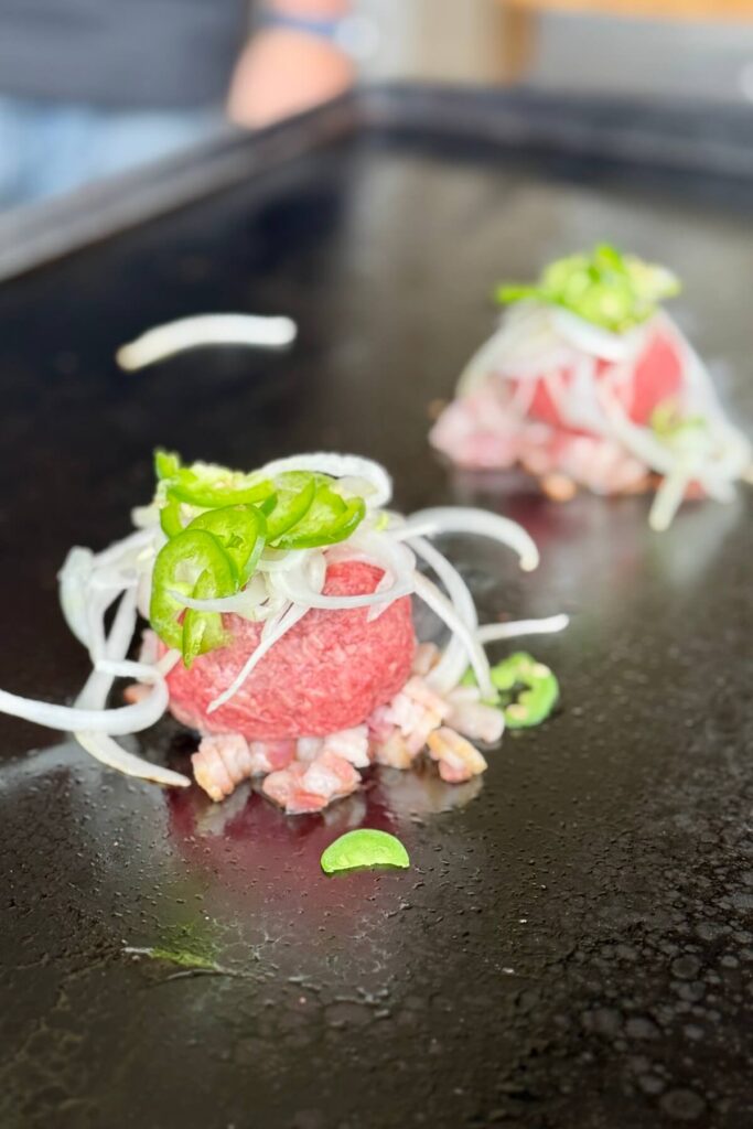 ground beef balls with bacon, sliced onion, and sliced jalapeños on a griddle