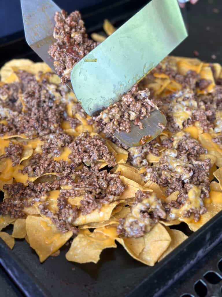 using a spatula to add ground beef and melted cheese to nacho chips on a griddle