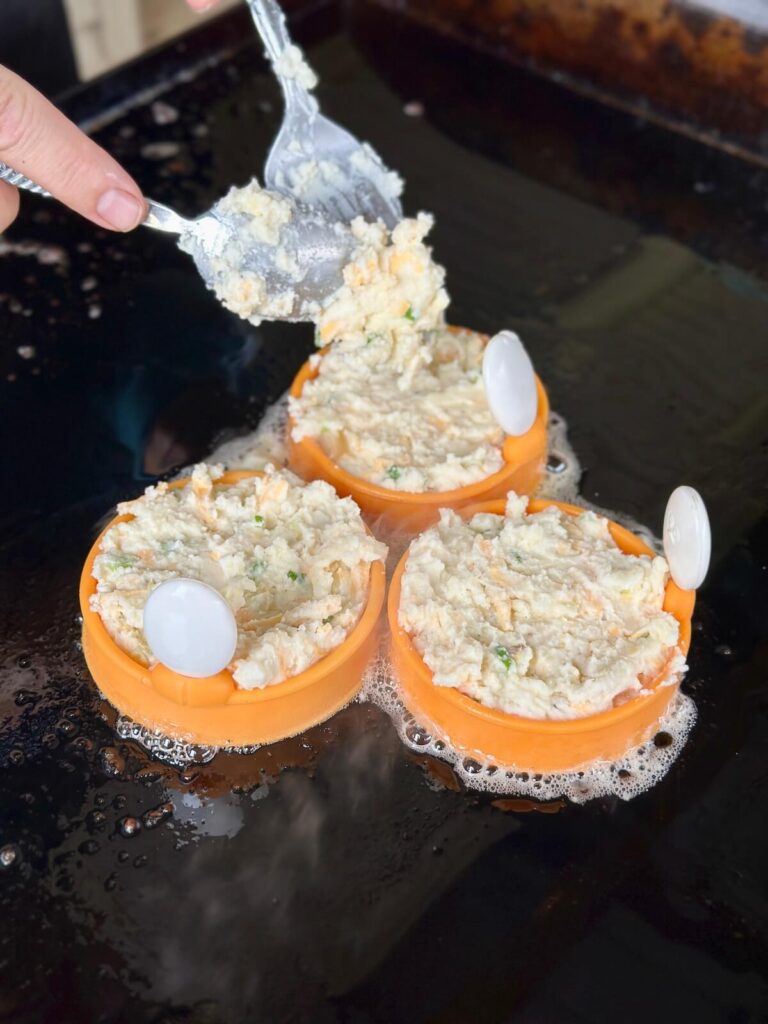 spooning cheesy leftover mashed potatoes mixture into egg molds on a griddle