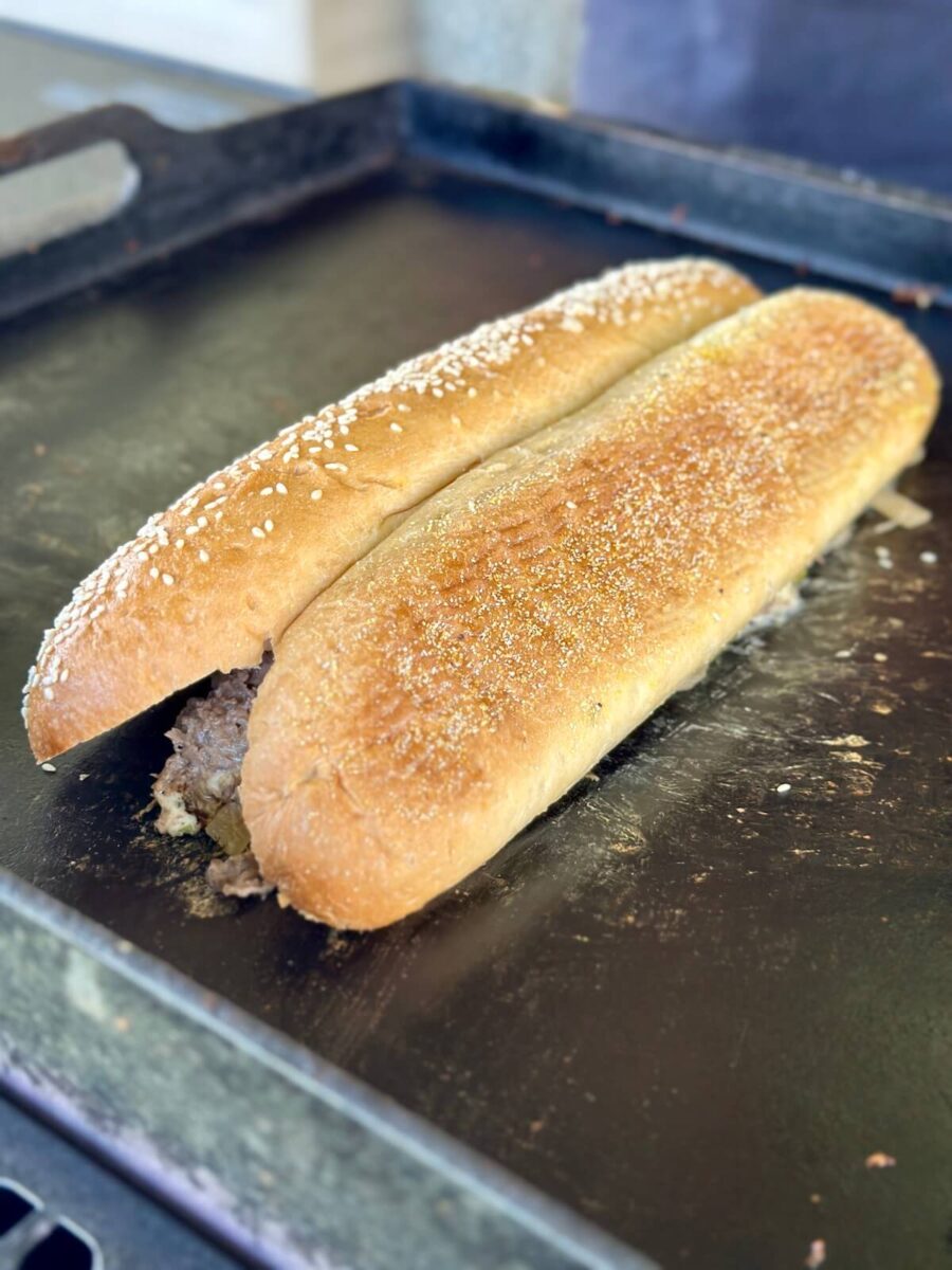 steaming a hoagie roll cut side down on top of a pile of cheesesteak mixture on a griddle