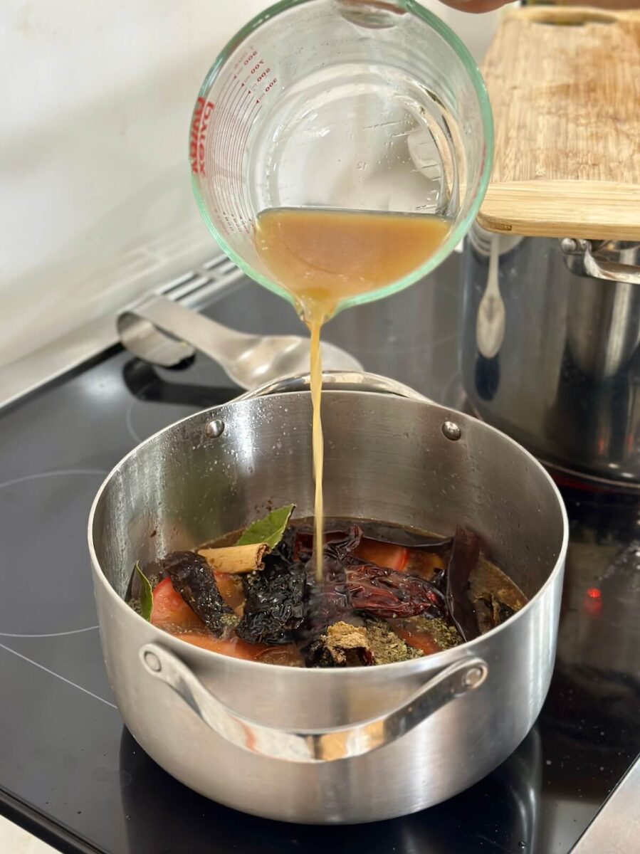 pouring beef stock in a pot with dried peppers, tomatoes, onions, and spices