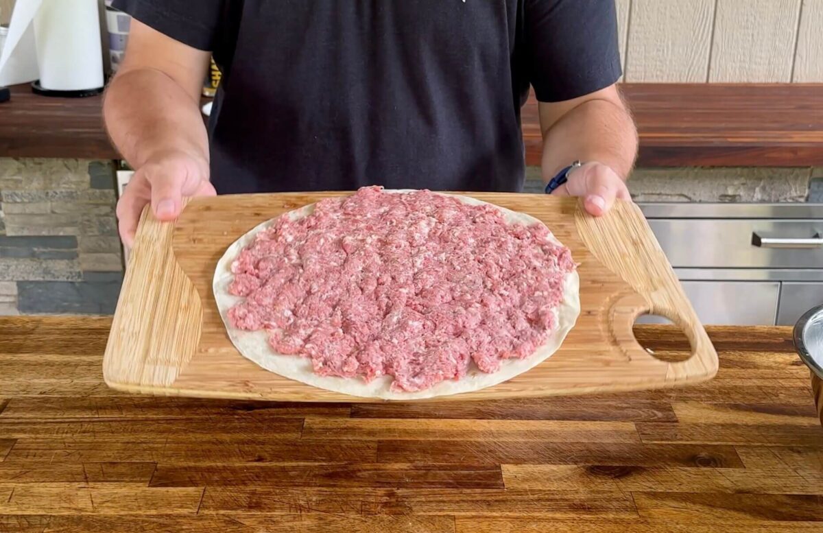 large flour tortilla with ground Italian sausage and beef in a thin even layer spread from edge to edge