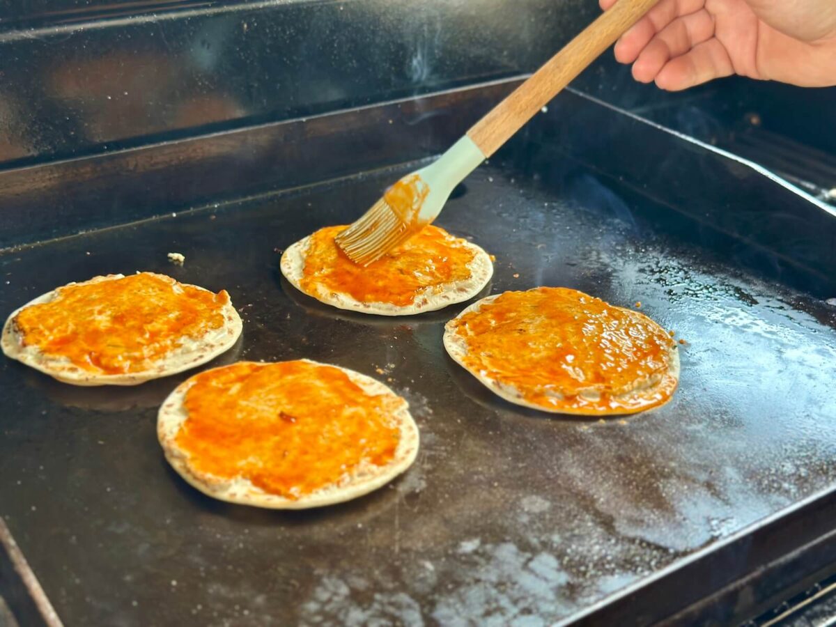 brushing buffalo sauce onto smashed chicken tacos on a griddle