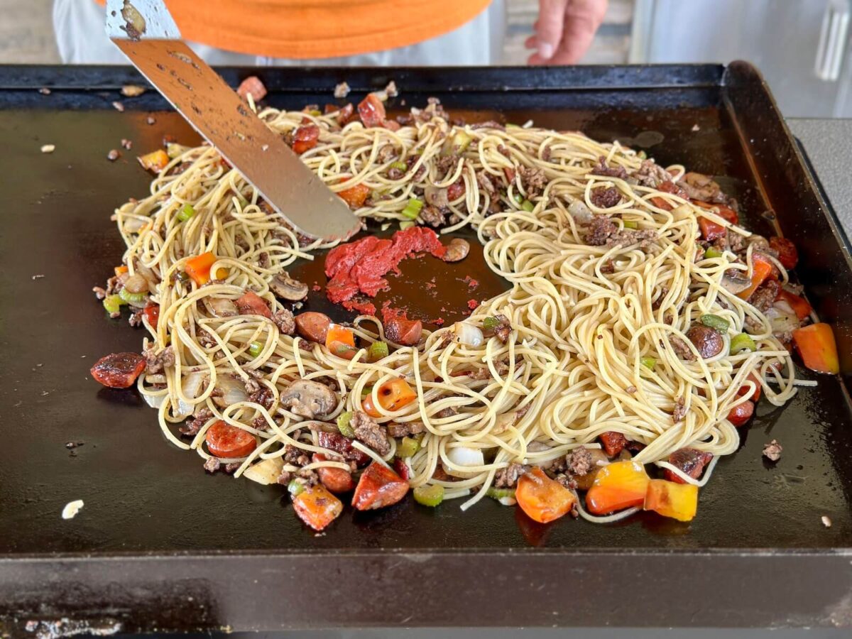 heating tomato paste in a well in the middle of a pile of spaghetti noodles with ground beef, sausage, and vegetables
