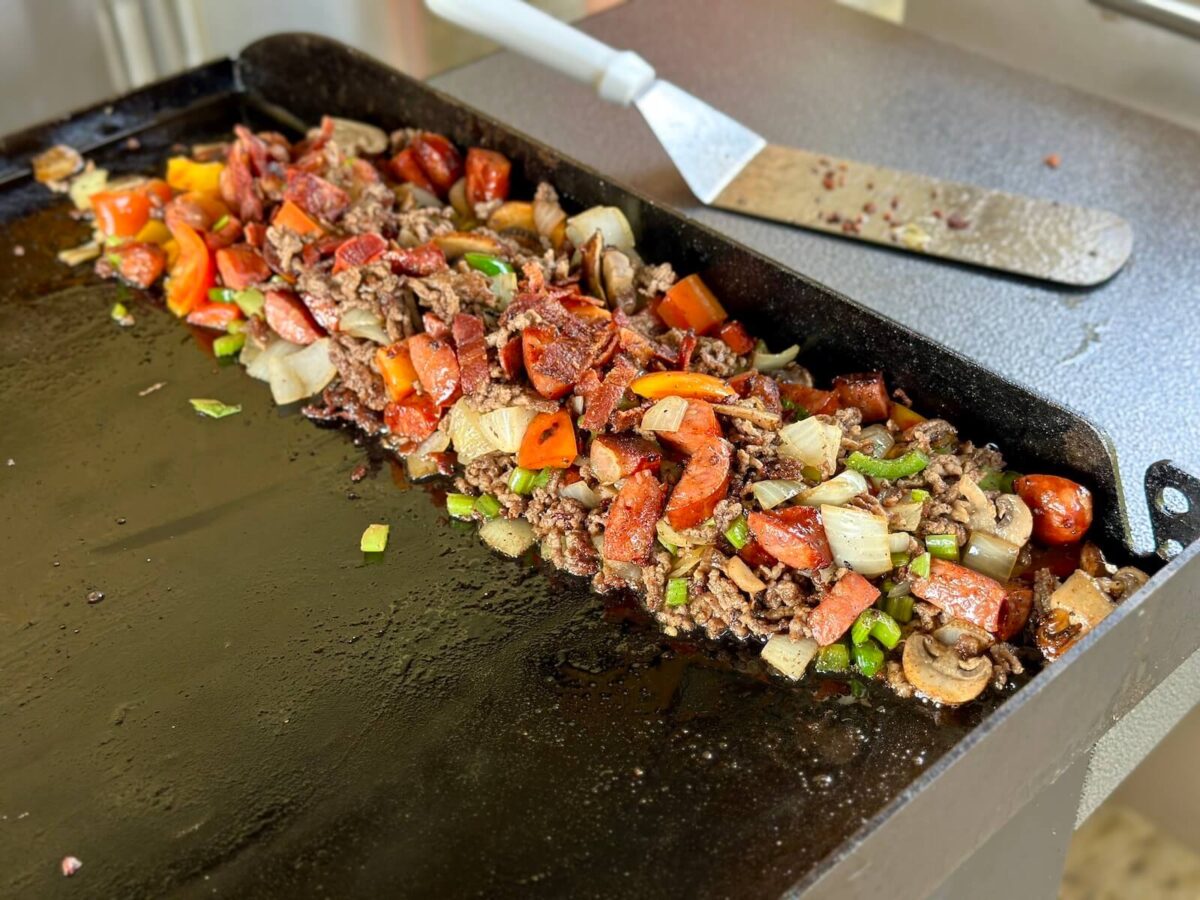 ground beef, andouille sausage, bacon, and vegetables mixture pushed to the side of a Blackstone griddle