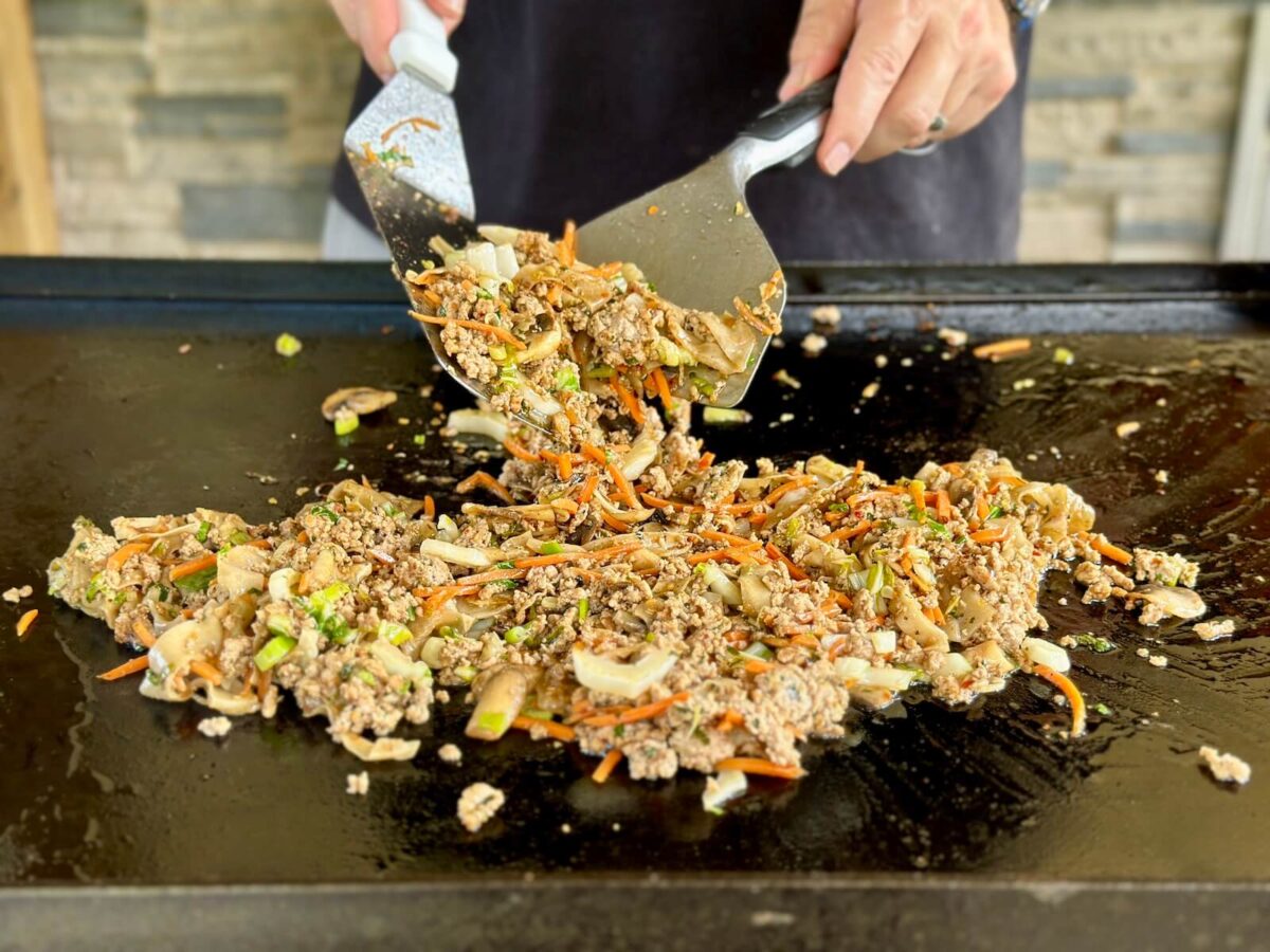 mixing ground pork, wonton noodles, shredded carrots, and other dumpling ingredients on a Blackstone griddle