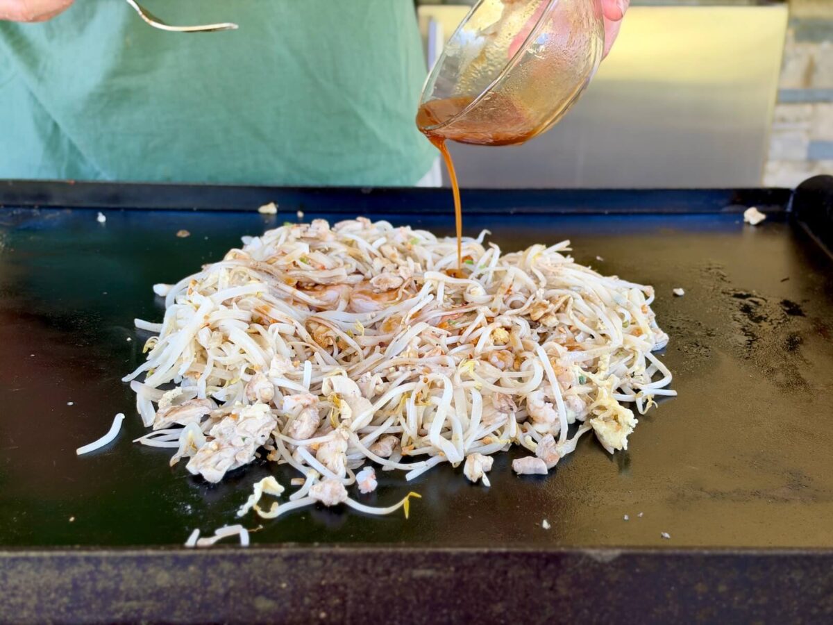 adding pad thai sauce to cooked noodles and other pad thai ingredients on a griddle