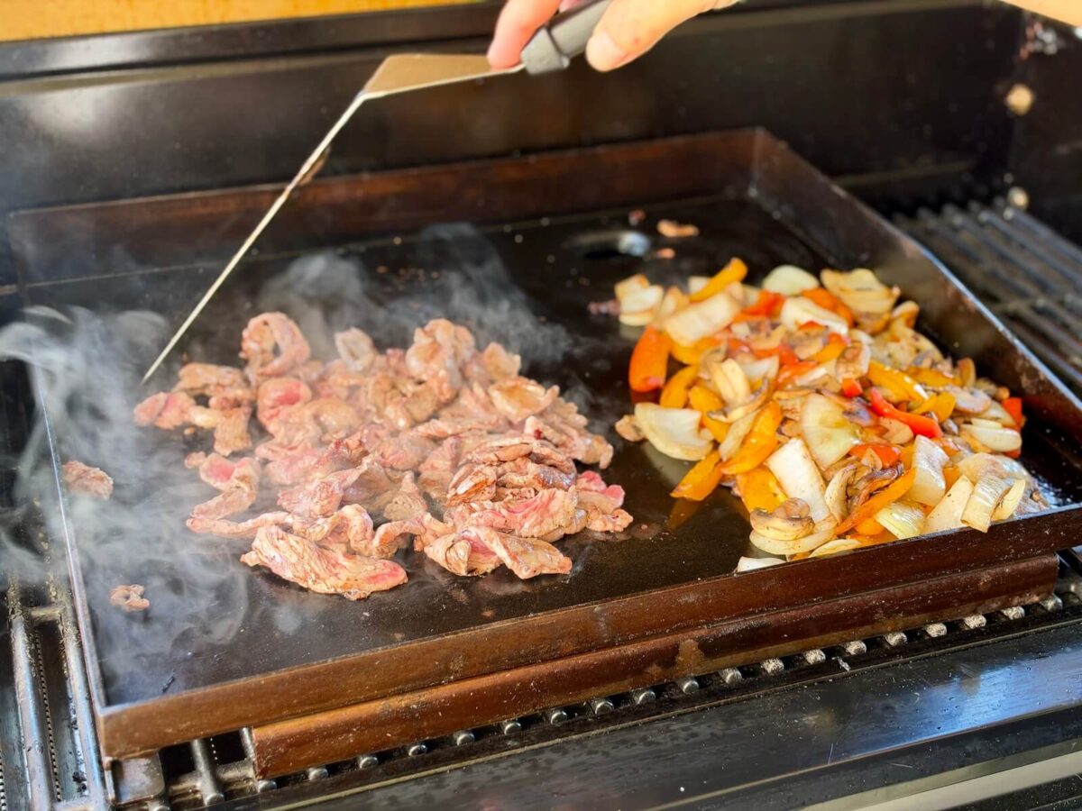 cooking sliced ribeye steak and chopped bell peppers and onions on a griddle attachment on a gas grill